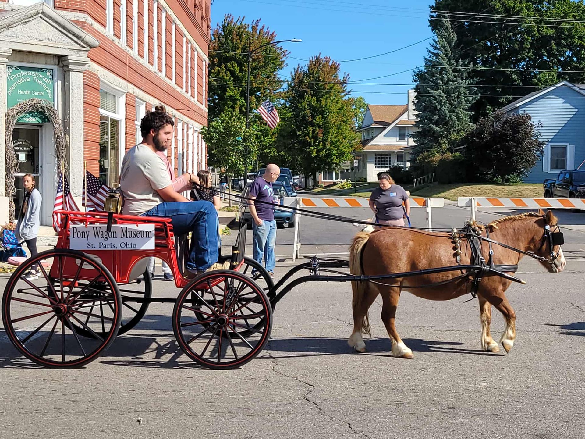 Man driving a horse-drawn cart down a street; red cart, brown horse, brick building in background.