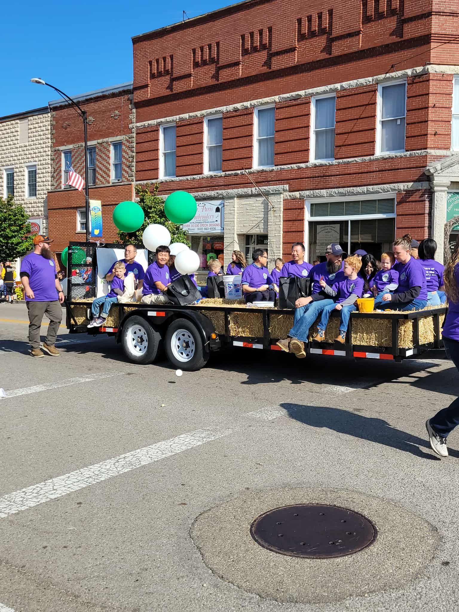Parade float with people in purple shirts, green and white balloons, and brick building background.