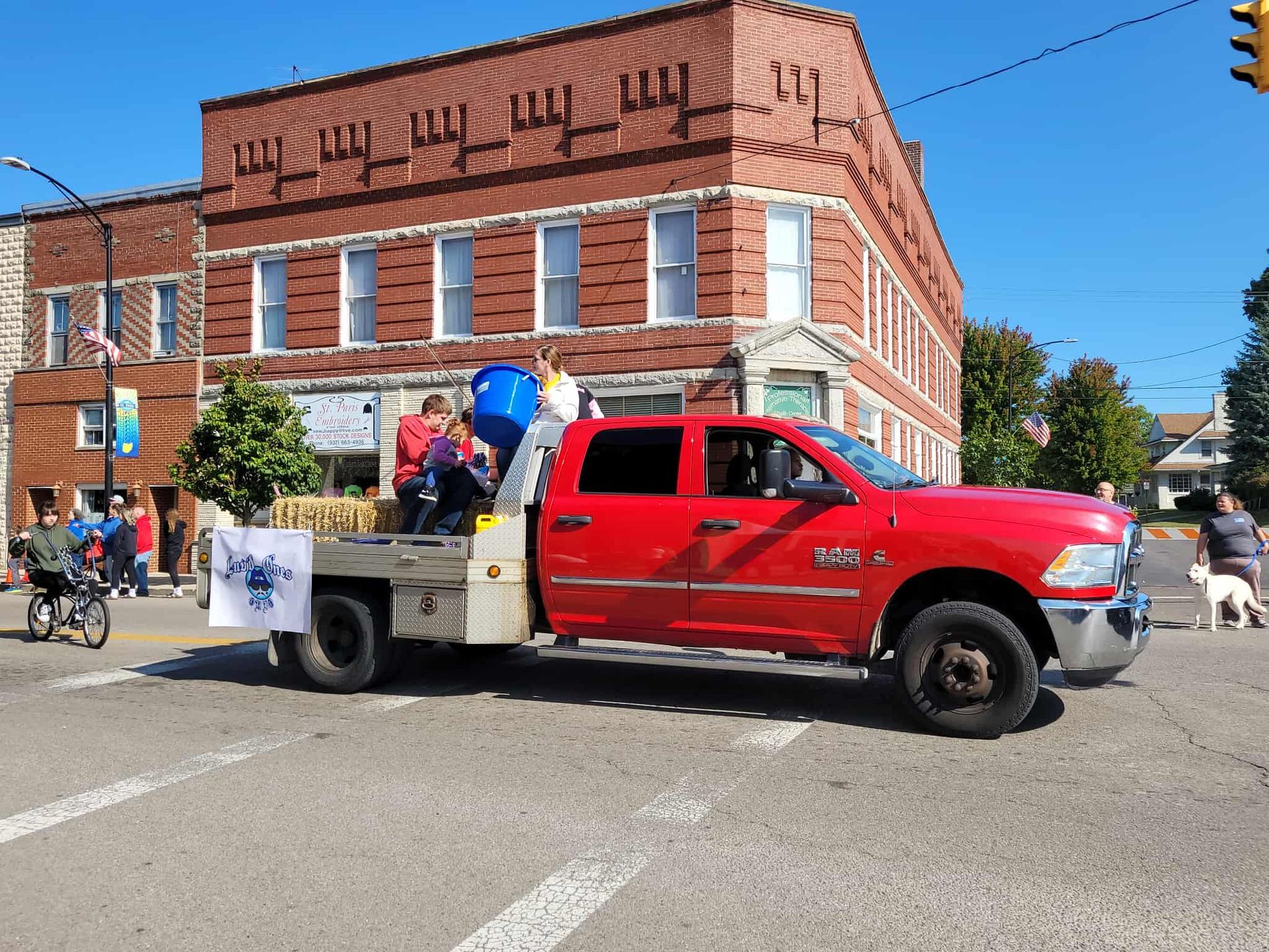 Red pickup truck carrying people in a parade, with a brick building in the background.