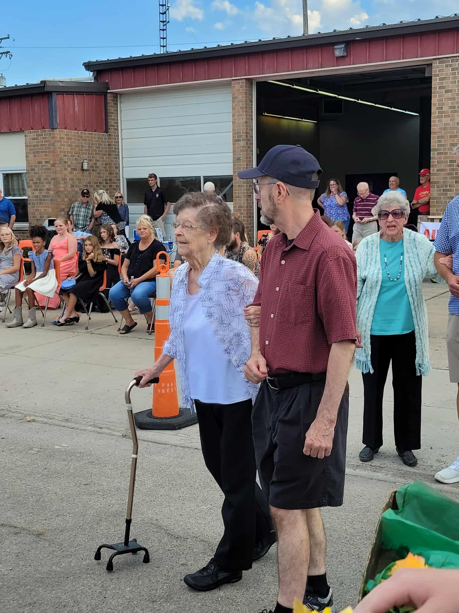 An older woman using a cane is helped by a man, walking near a brick building; many people watch.