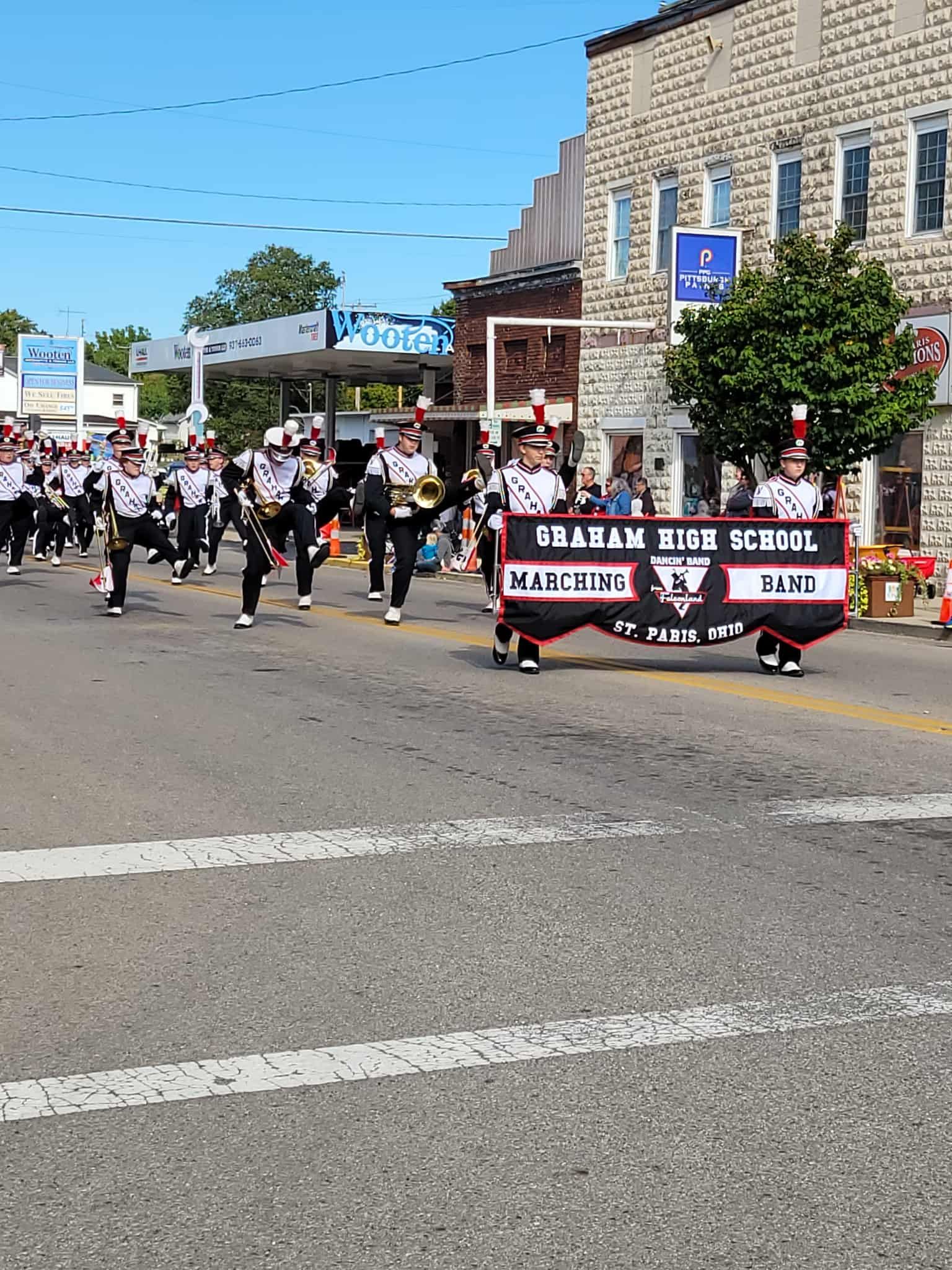 Marching band in black and white uniforms marches down street, carrying banner. Buildings in background.