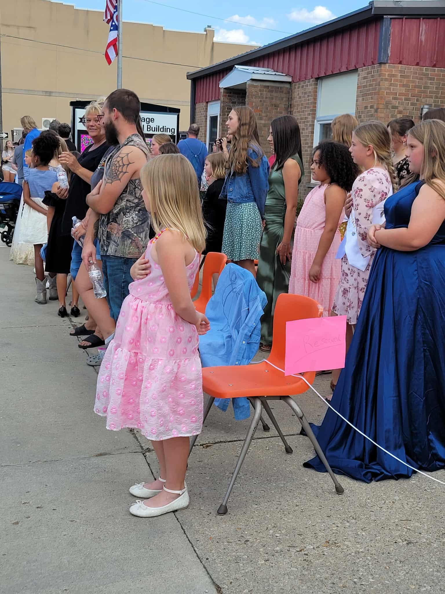 People standing in a line outdoors; a girl in a pink dress in front.