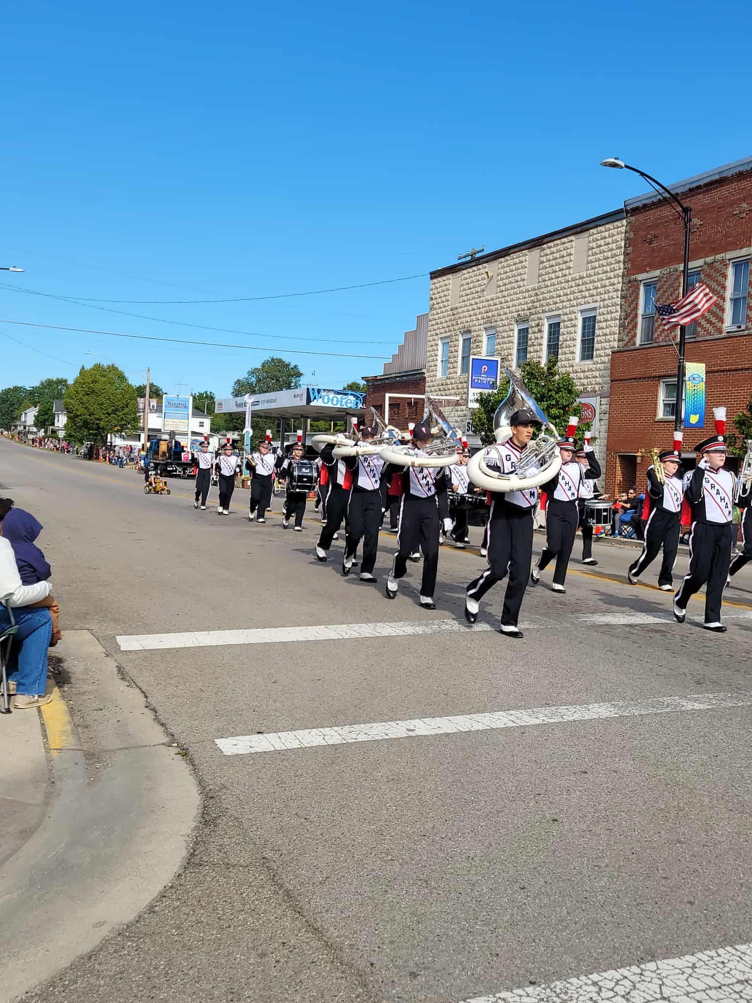 Marching band in black and white uniforms, playing music down a town street with buildings and blue sky.