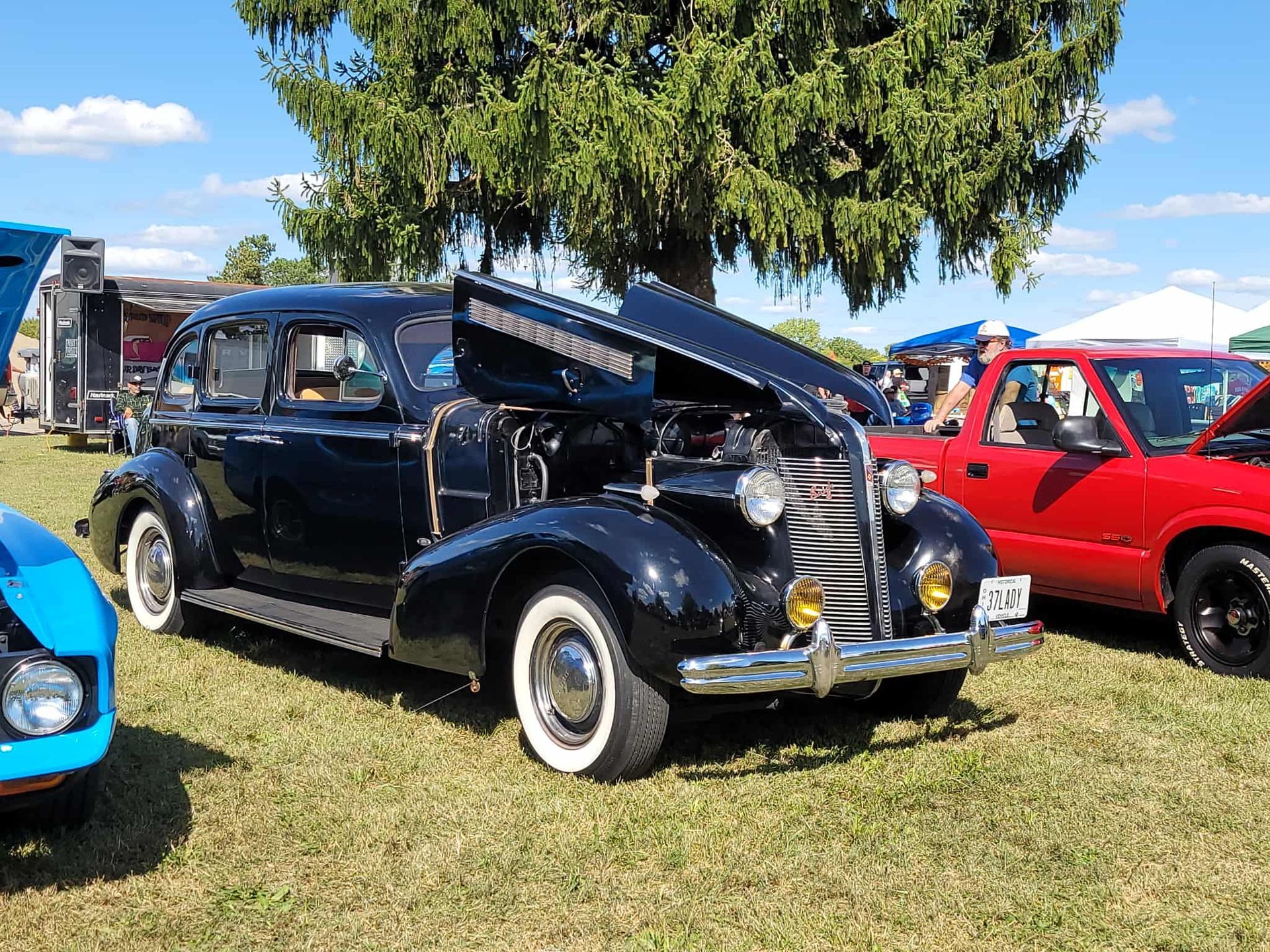 Black classic car with hood open, parked on grass at an outdoor car show, with a red truck visible.