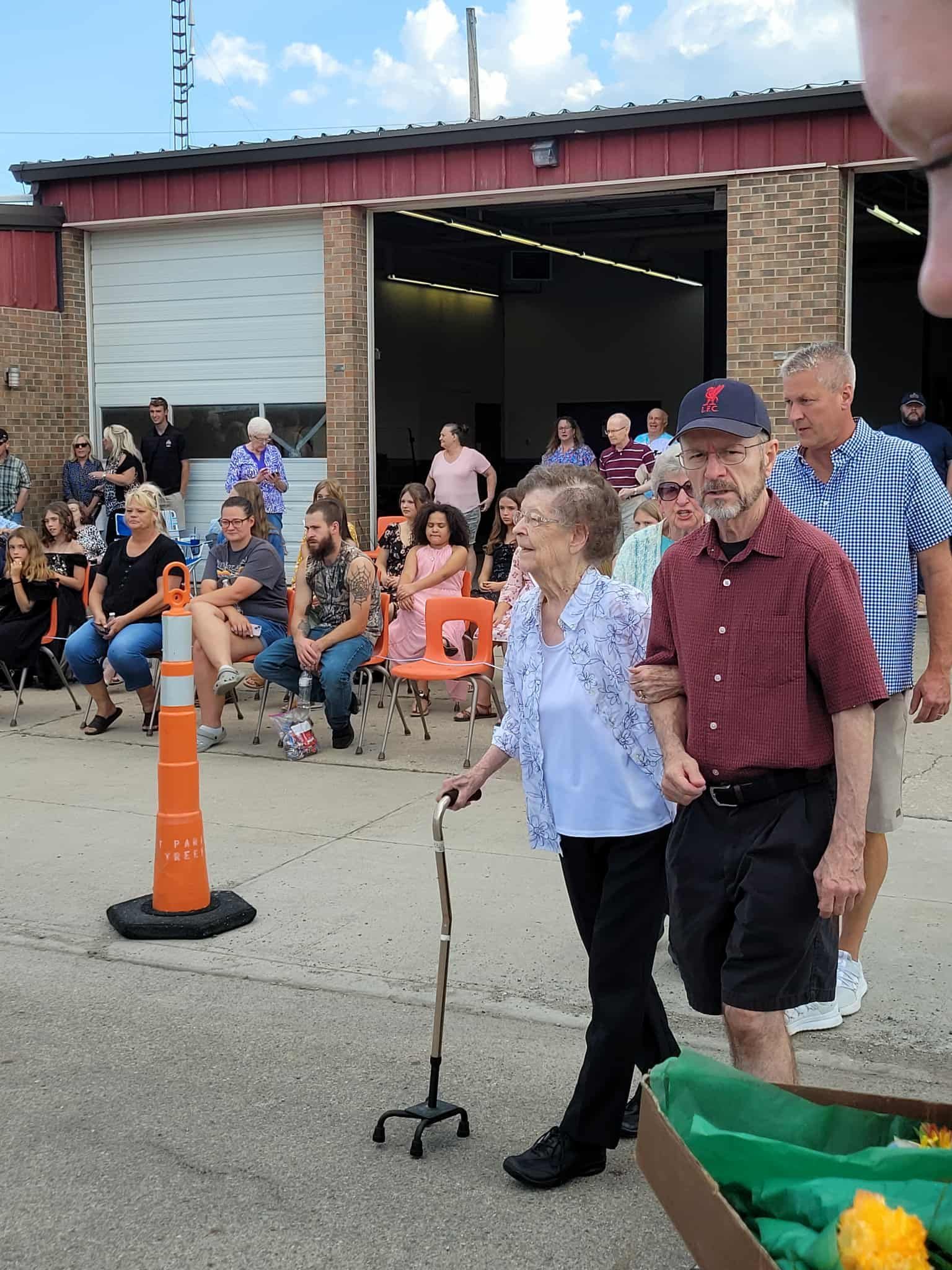 Elderly couple walking, man assisting woman with cane; fire station background. Crowd of people watching.
