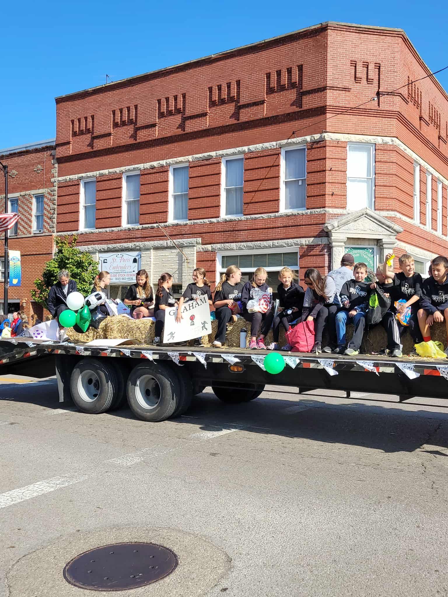 Children on a parade float in front of a brick building with a blue sky.
