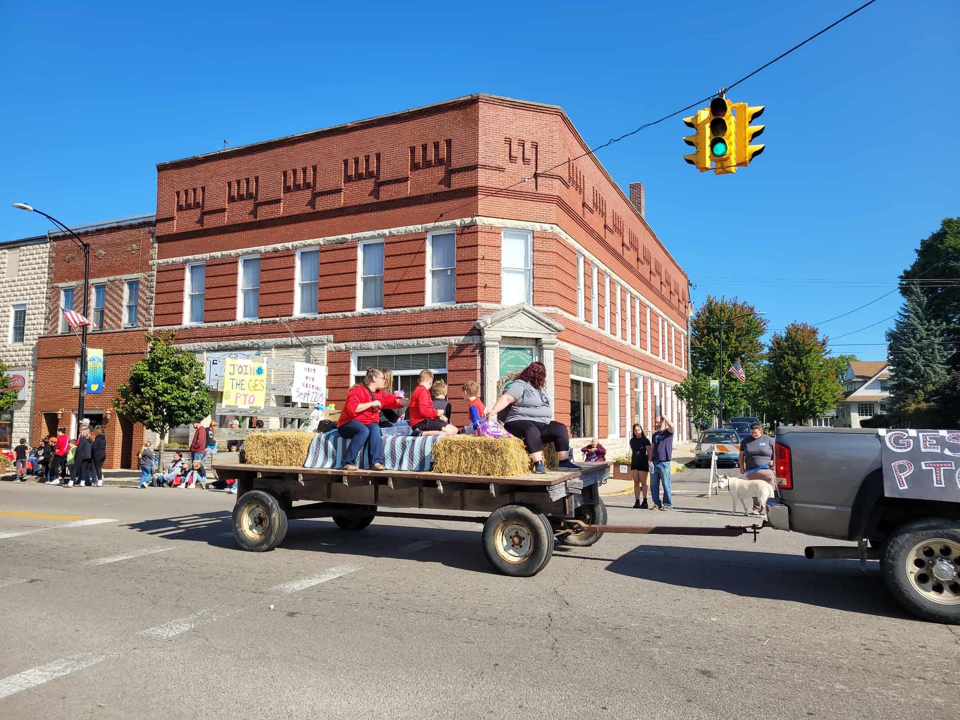 People on hay bale float in a parade, brick building in background, green traffic light overhead.