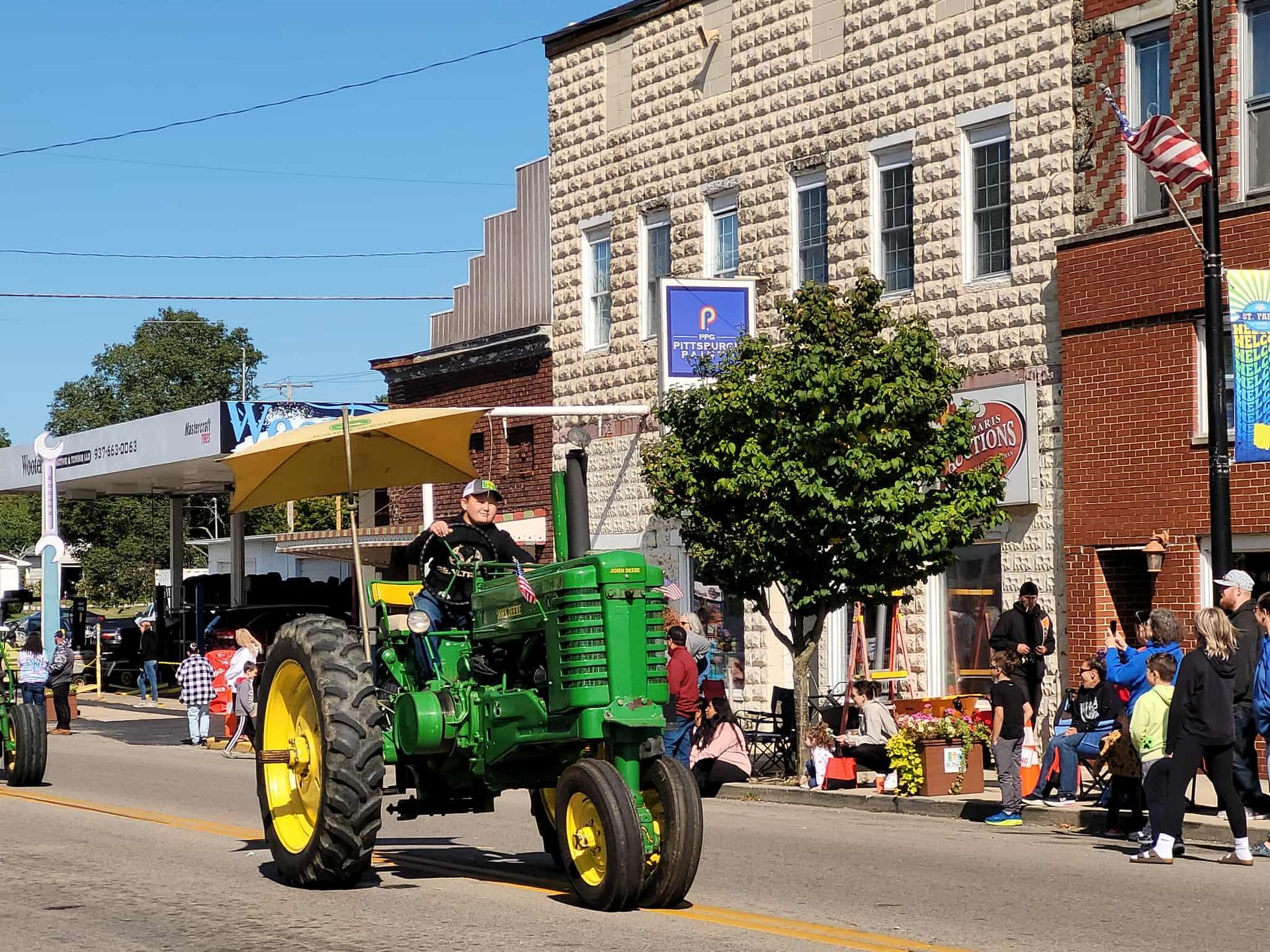 Green John Deere tractor in parade, downtown street. People watching from sidewalk.