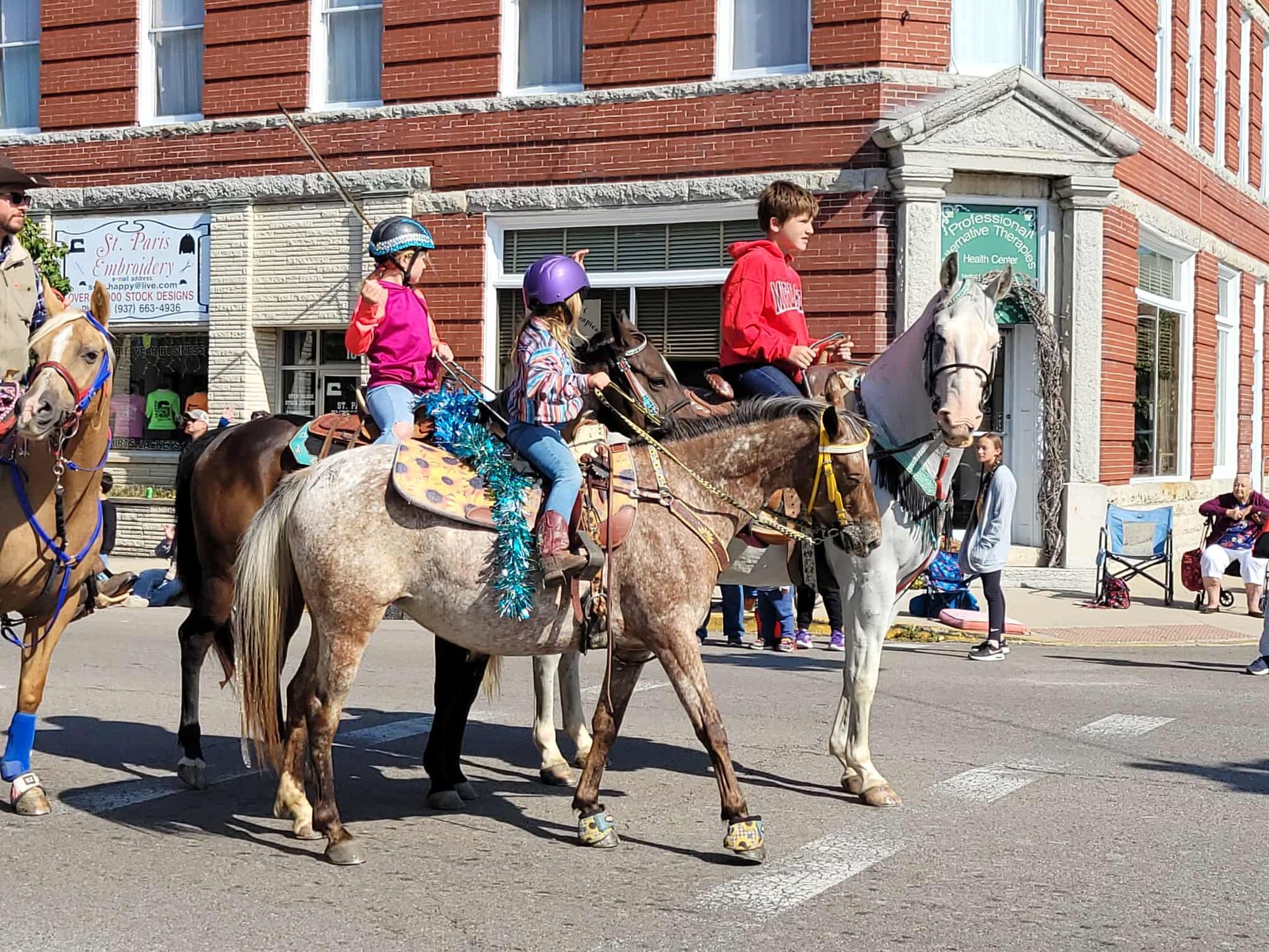 Children on horses in a parade, riding past a brick building.