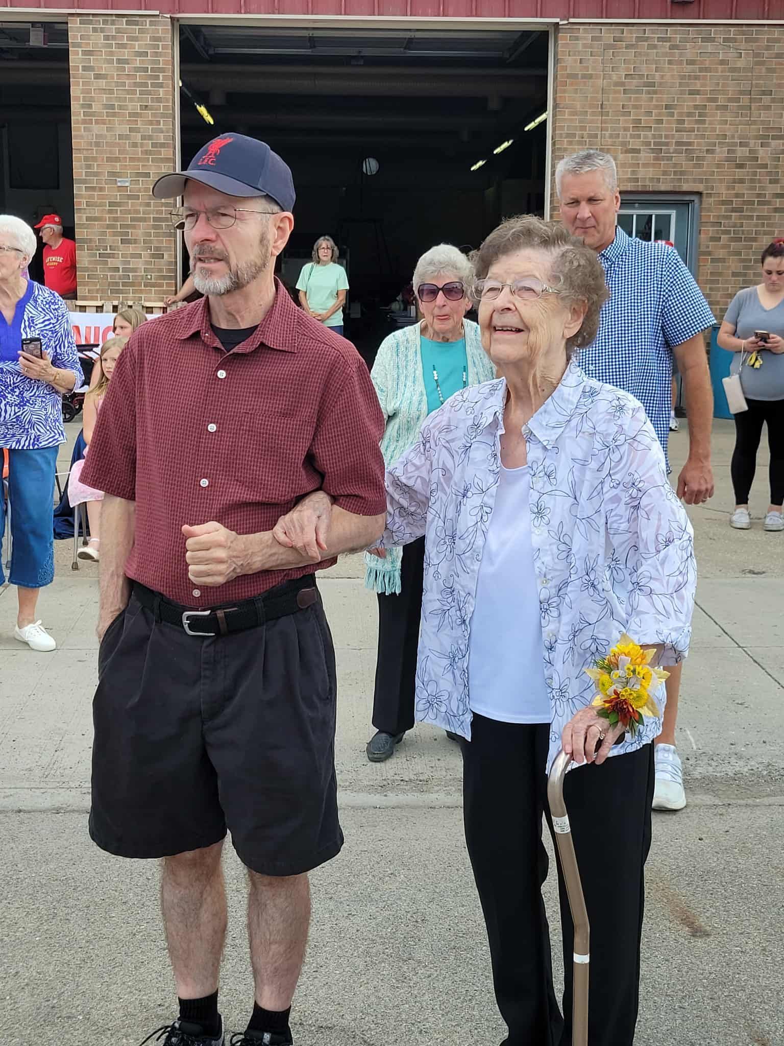 Man assists woman walking, arm in arm. Woman holds cane, smiles, and wears floral top. Others watch.