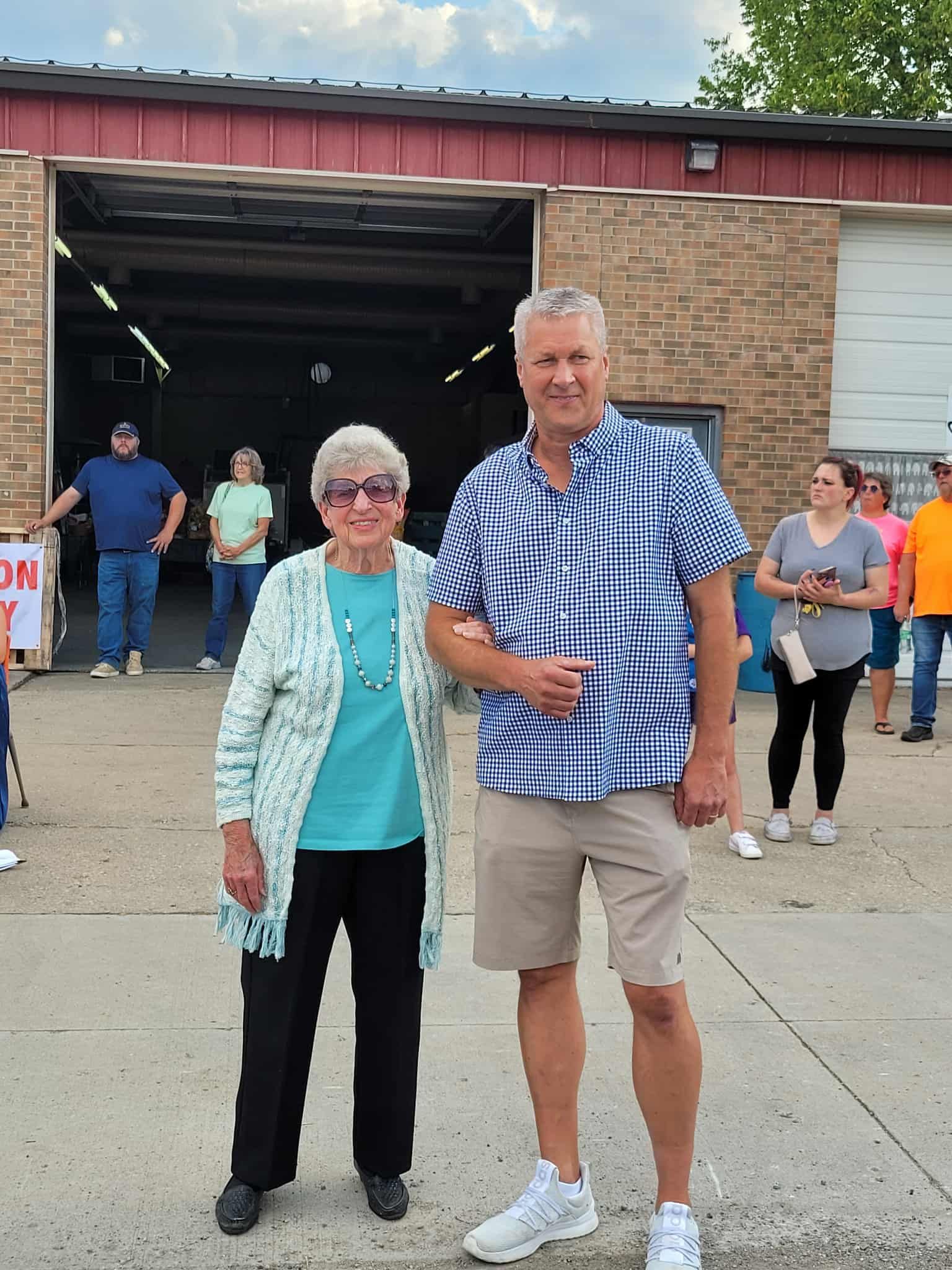 Man and woman stand in front of a building. Man wears a blue shirt, khaki shorts. Woman has a cardigan, glasses. People behind them.