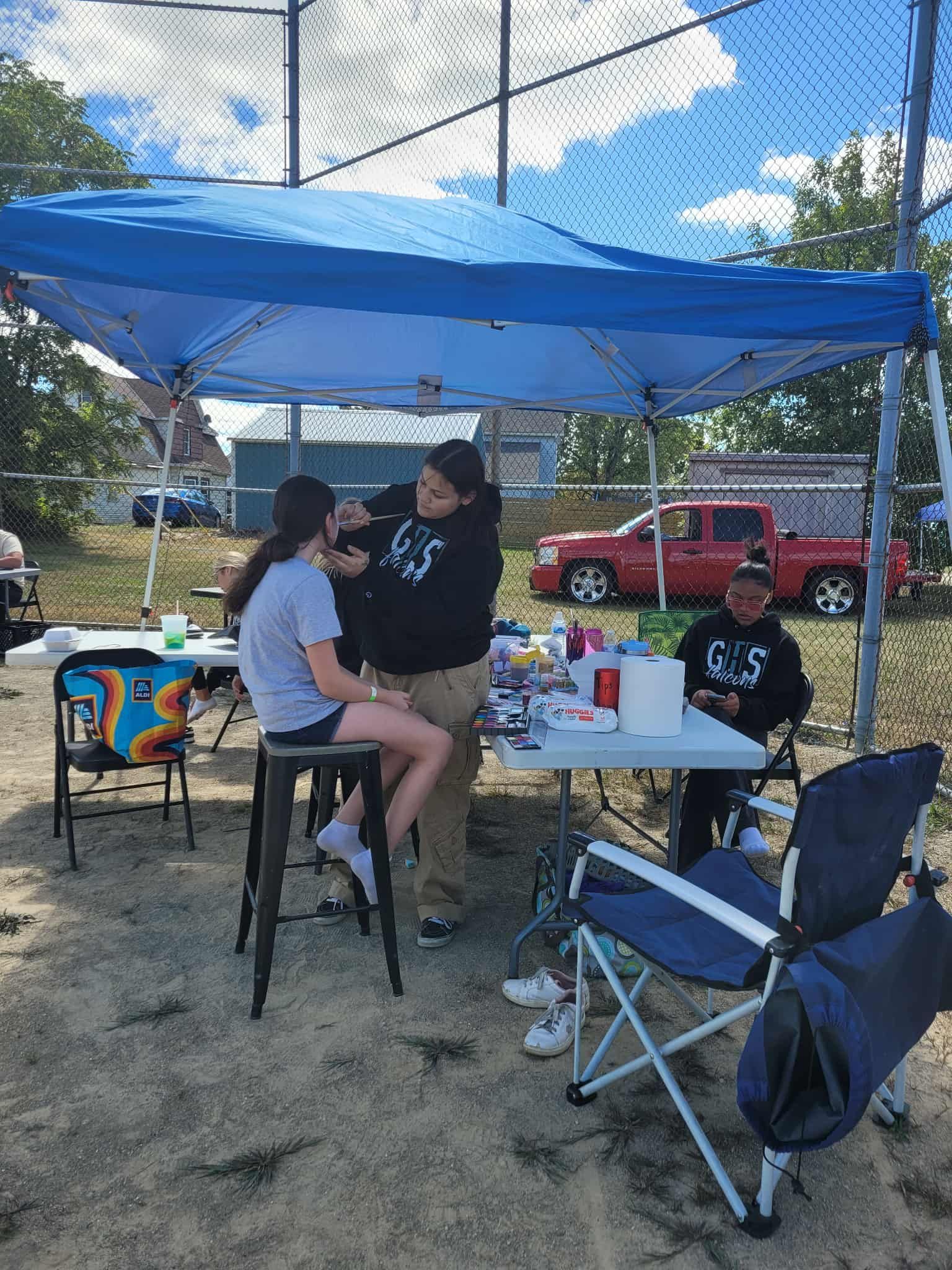 Person getting face painted under a blue canopy. Another person sits, a red truck in background.