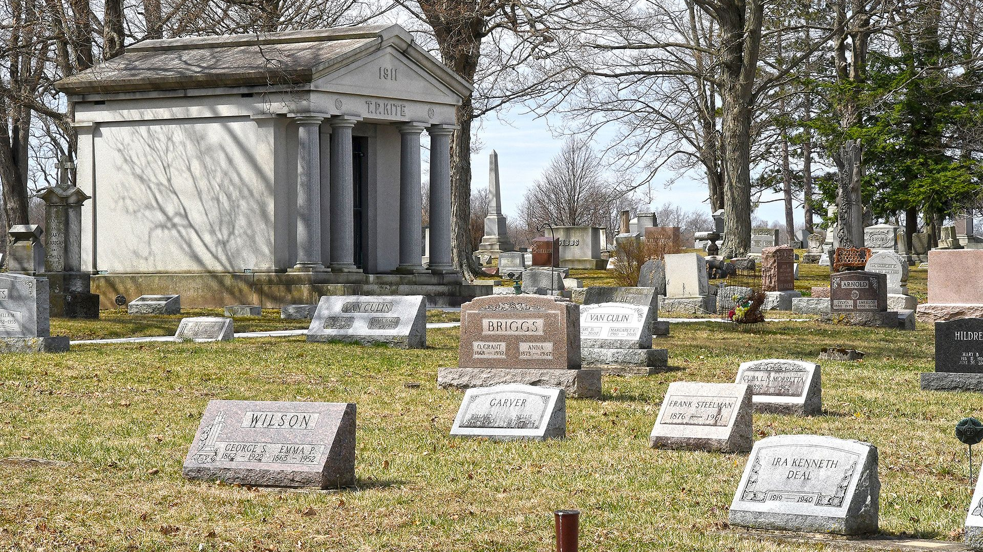 Cemetery scene with headstones and a classical mausoleum under a cloudy sky.