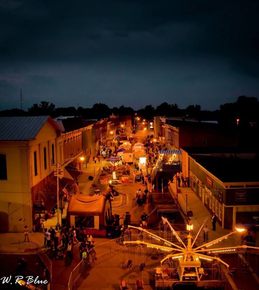 Nighttime street fair, crowds, lit buildings, Ferris wheel, vendor stalls.