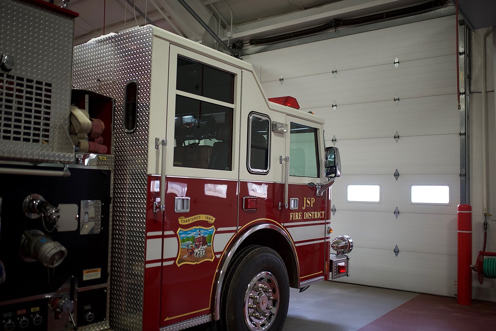 Fire truck parked in a fire station bay. Red and white paint, overhead garage door visible.