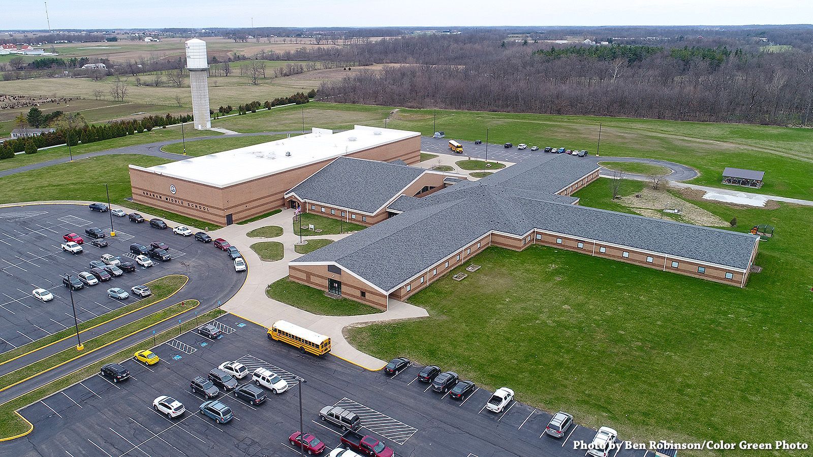 Aerial view of a school with parking lot, water tower in background. Green grass and trees surround the building.
