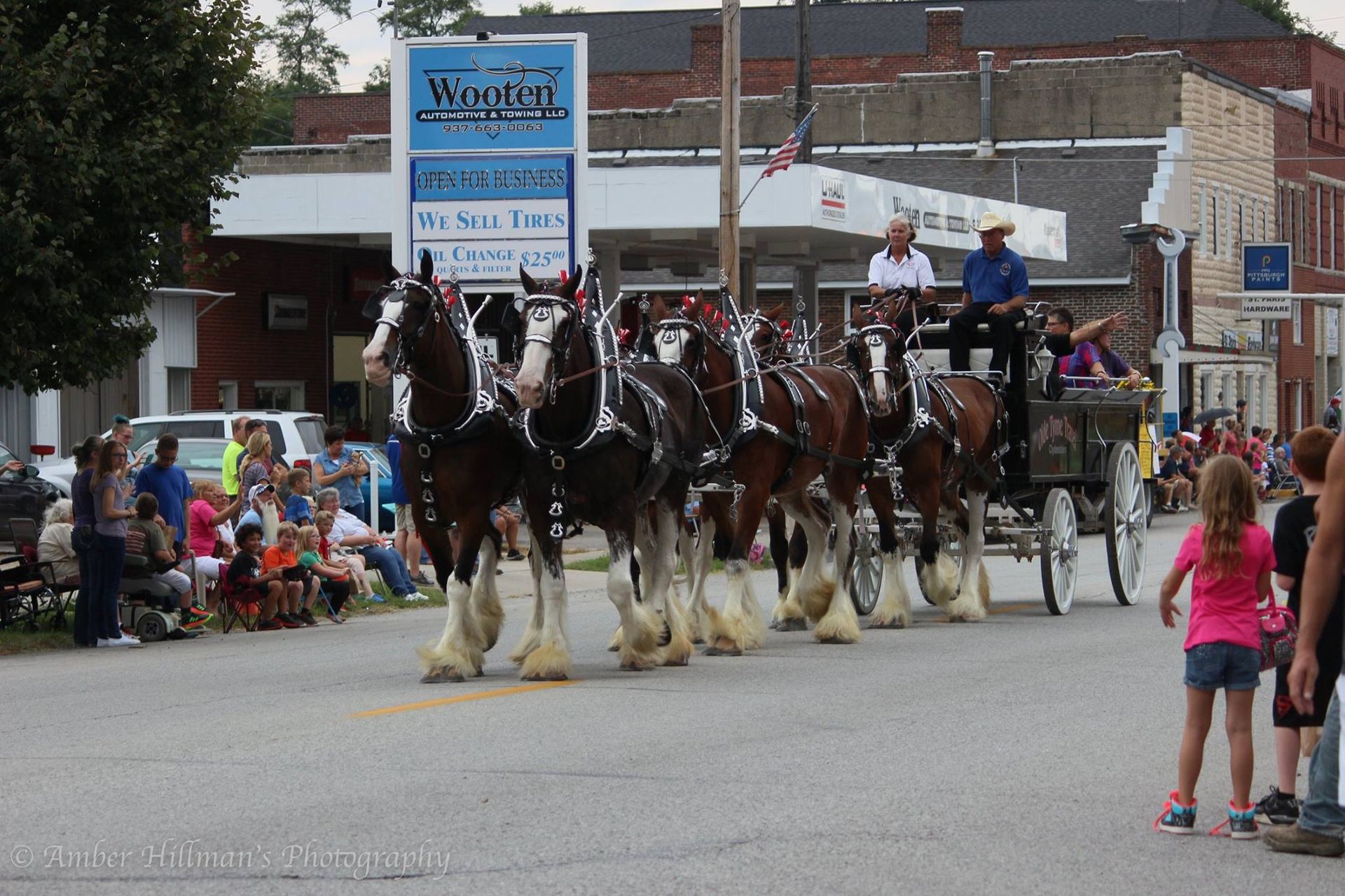 Eight Clydesdale horses pulling a wagon in a parade; spectators watch from the roadside.