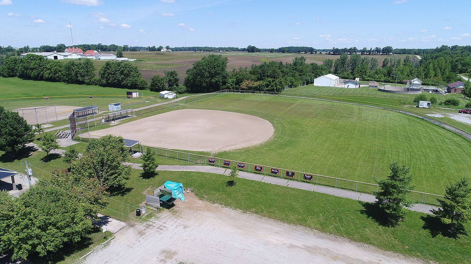 Aerial view of a baseball field and surrounding park with green grass, trees, and buildings on a sunny day.