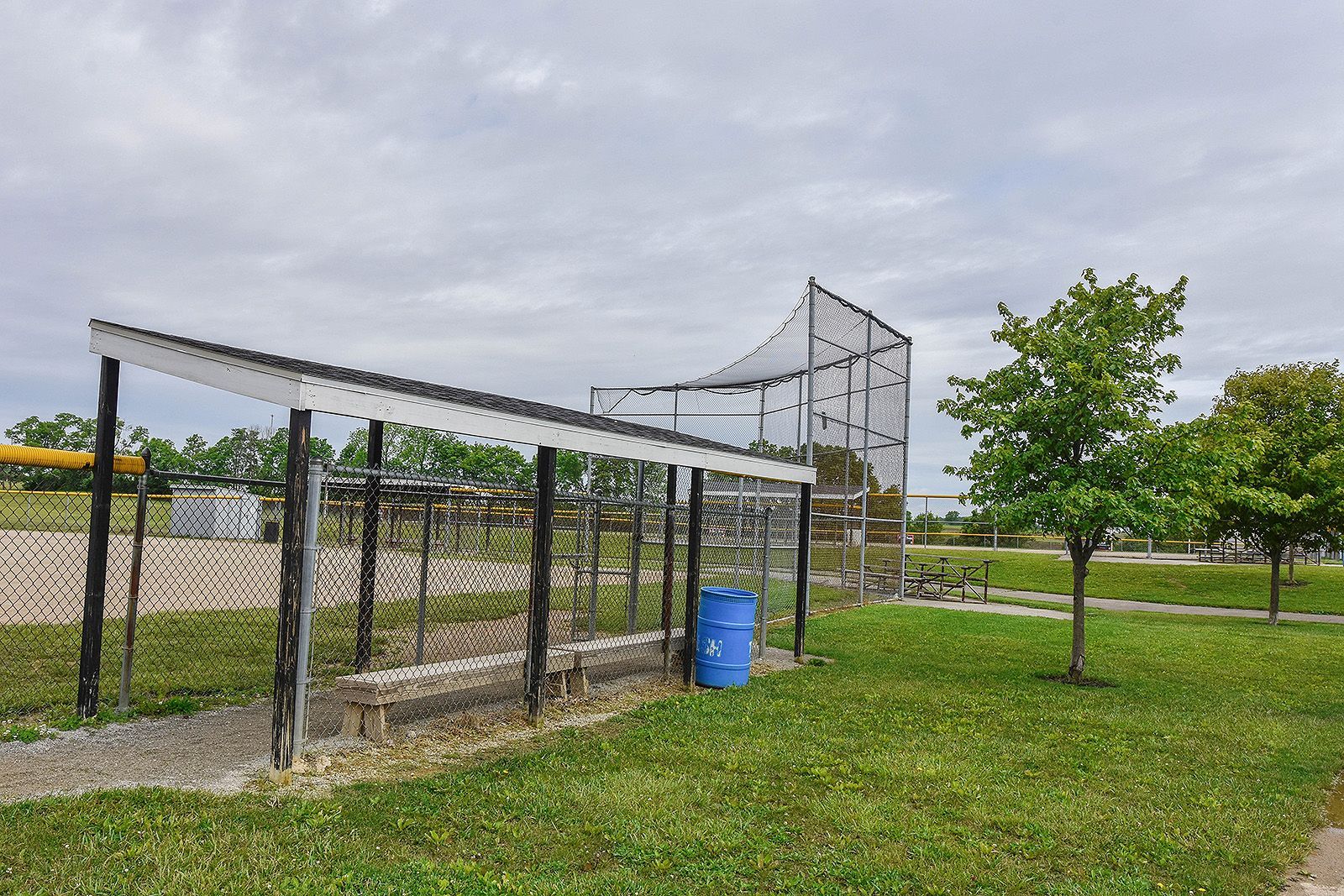 Baseball batting cage with roof and netting. A blue barrel sits on the grass next to a gravel path and small trees.