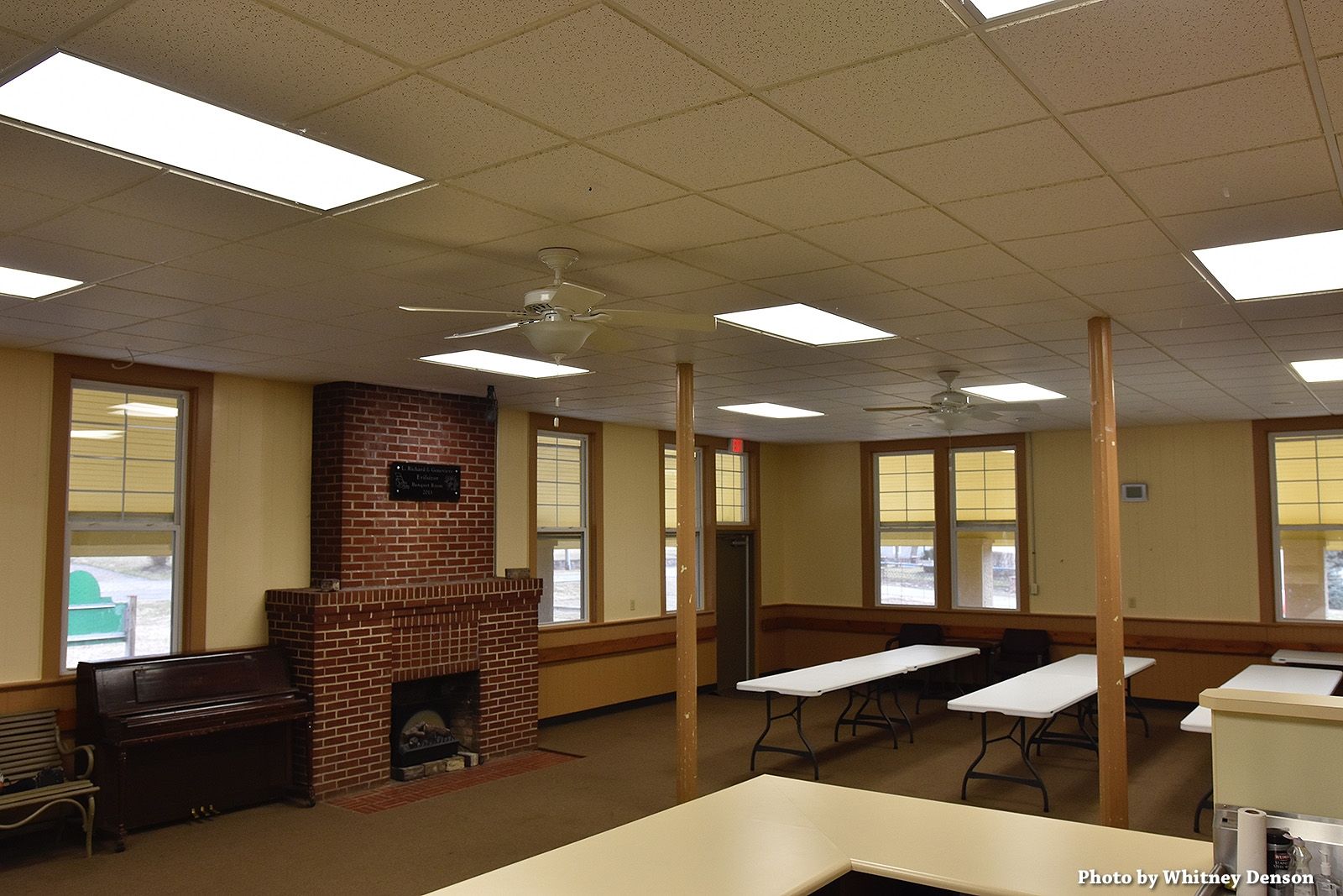 Interior of a room with a brick fireplace, tables, windows, and ceiling lights.