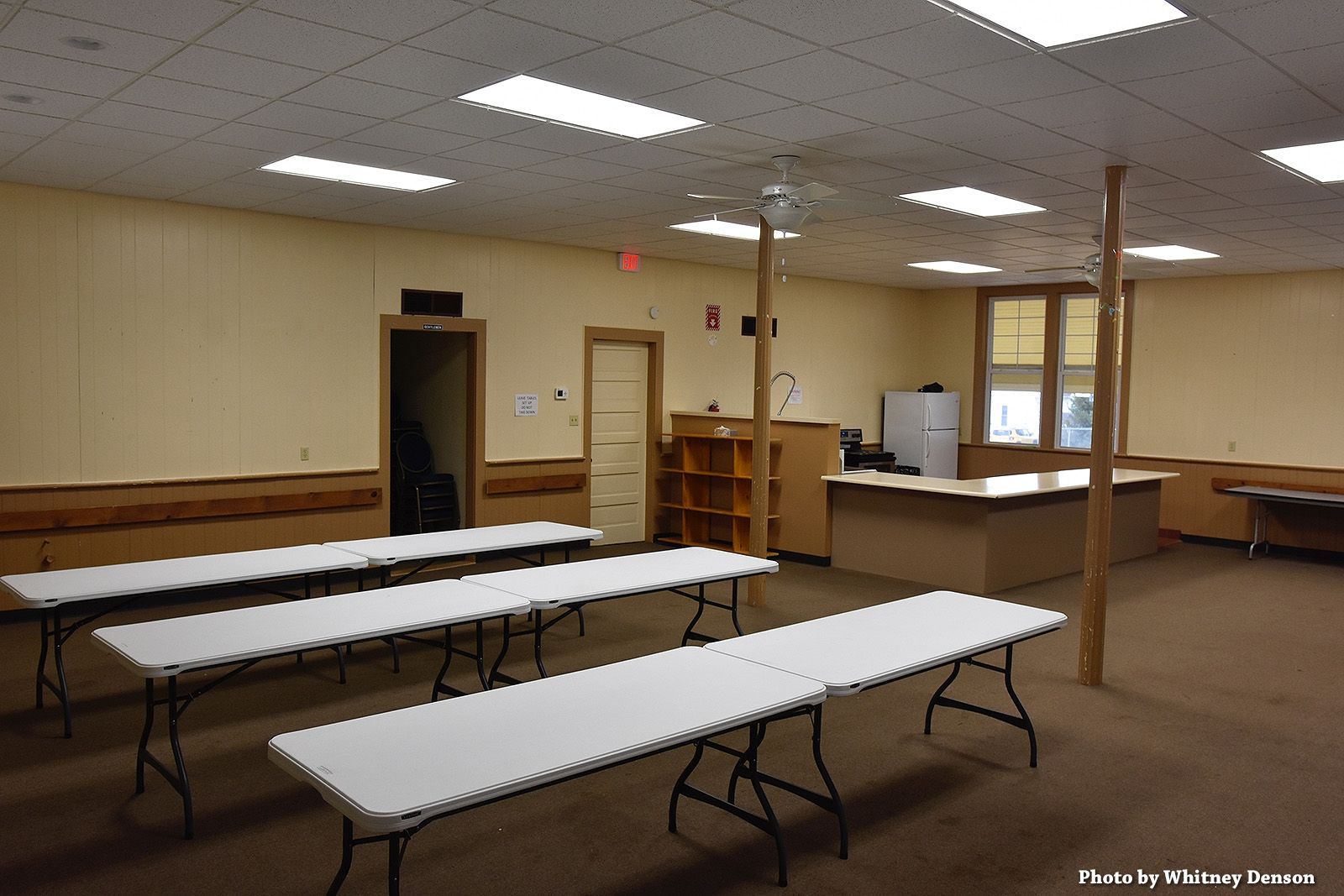An empty room with long white tables set up for an event, beige walls, and wood support beams.