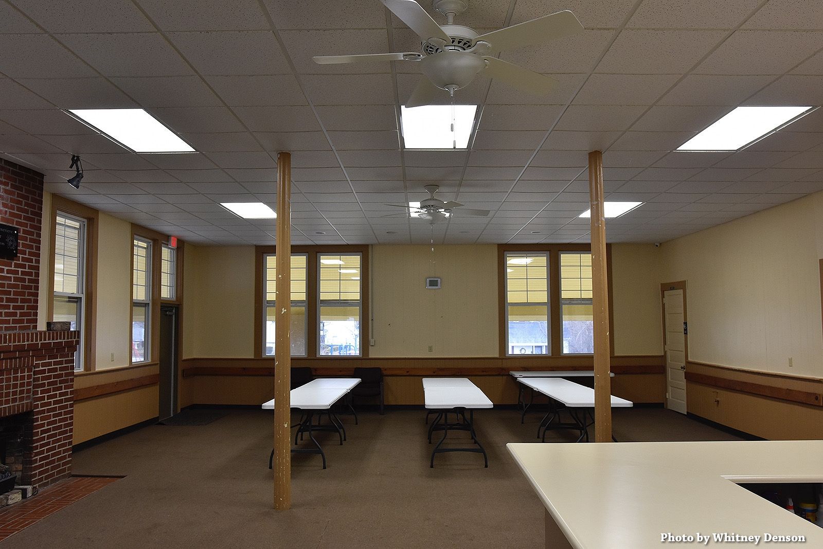 Empty community hall with tables, windows, ceiling lights, and two support beams.