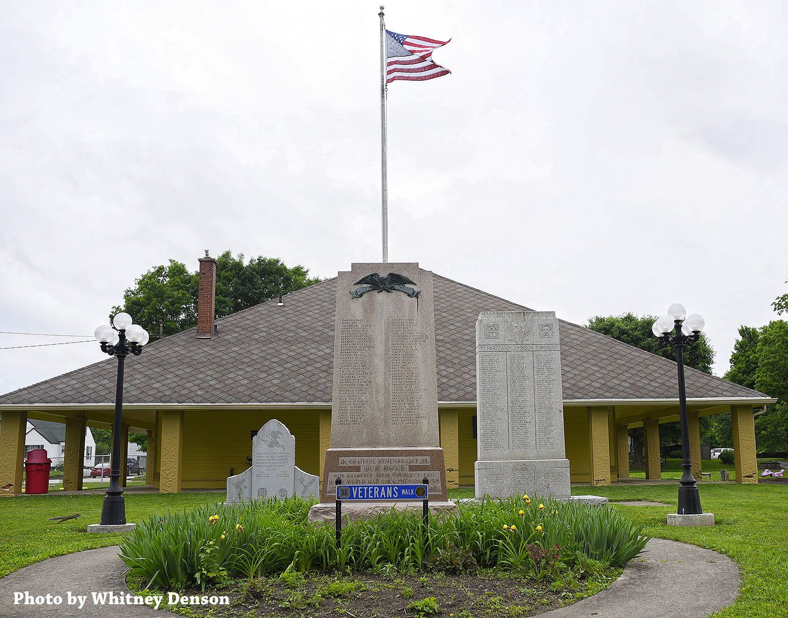 War memorial with American flag, names inscribed on stone, yellow building background.