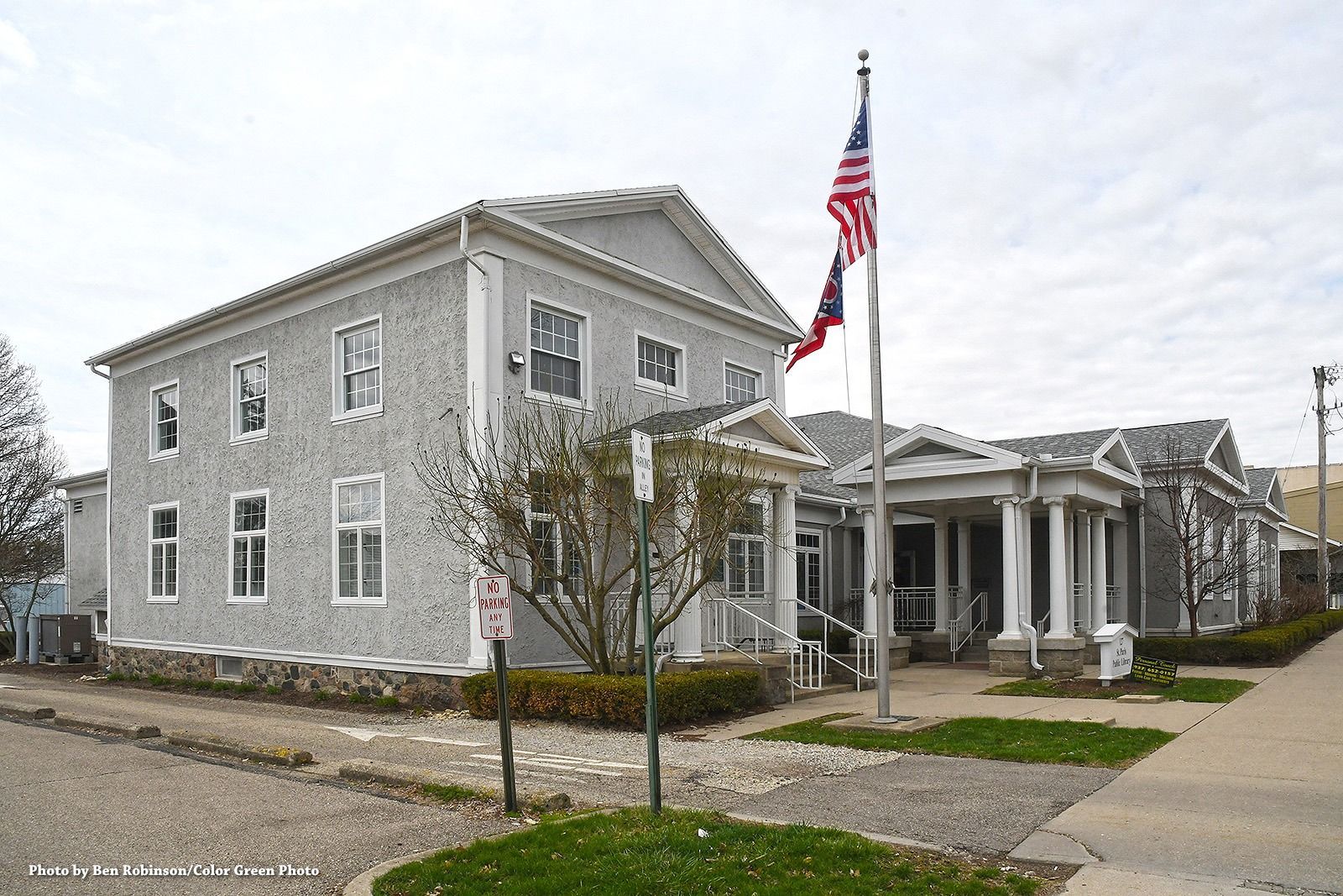 Two-story gray building with an American flag; a smaller building with columns and a sidewalk.