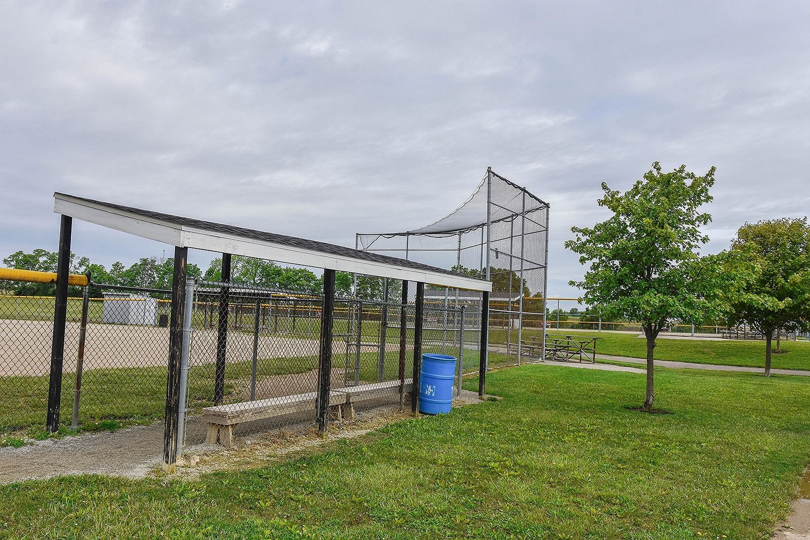 Baseball batting cage in a park. Green grass, blue barrel, netting, and cloudy sky.