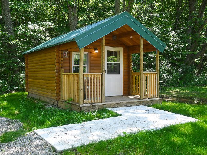 Small wooden cabin with green roof and porch in a wooded area.