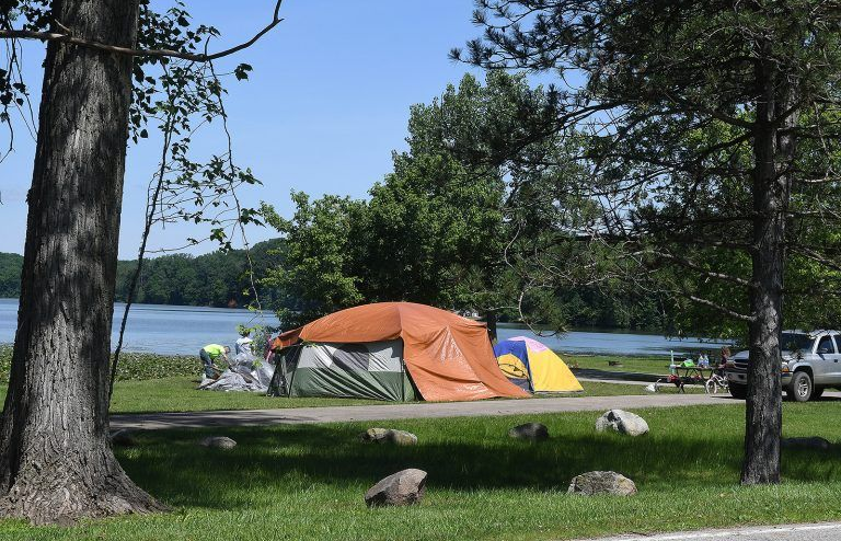 Campsite with two tents, near a lake. Trees in the foreground, with people setting up camp. Blue sky.