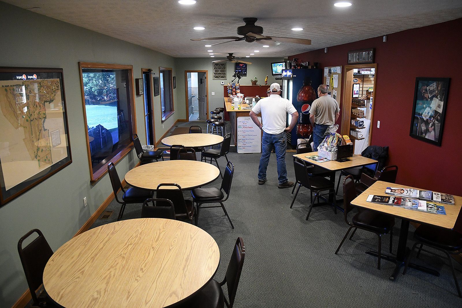 Interior of a small cafe with tables, chairs, and customers. Dark chairs and wooden tables; one person by counter.