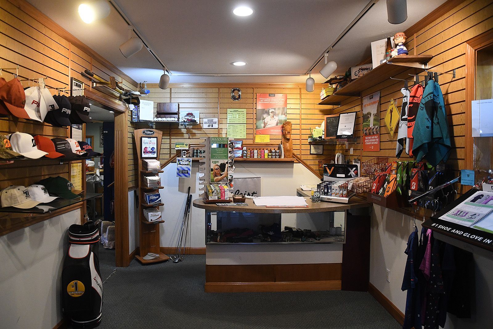 Golf shop interior with wooden shelves displaying hats, clubs, and accessories. A cash register sits at the counter.