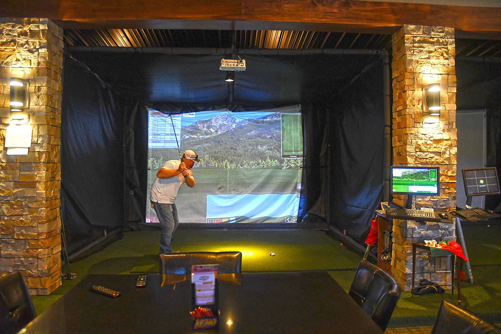 Man swings golf club in indoor golf simulator. Dark room with screen display and green turf.
