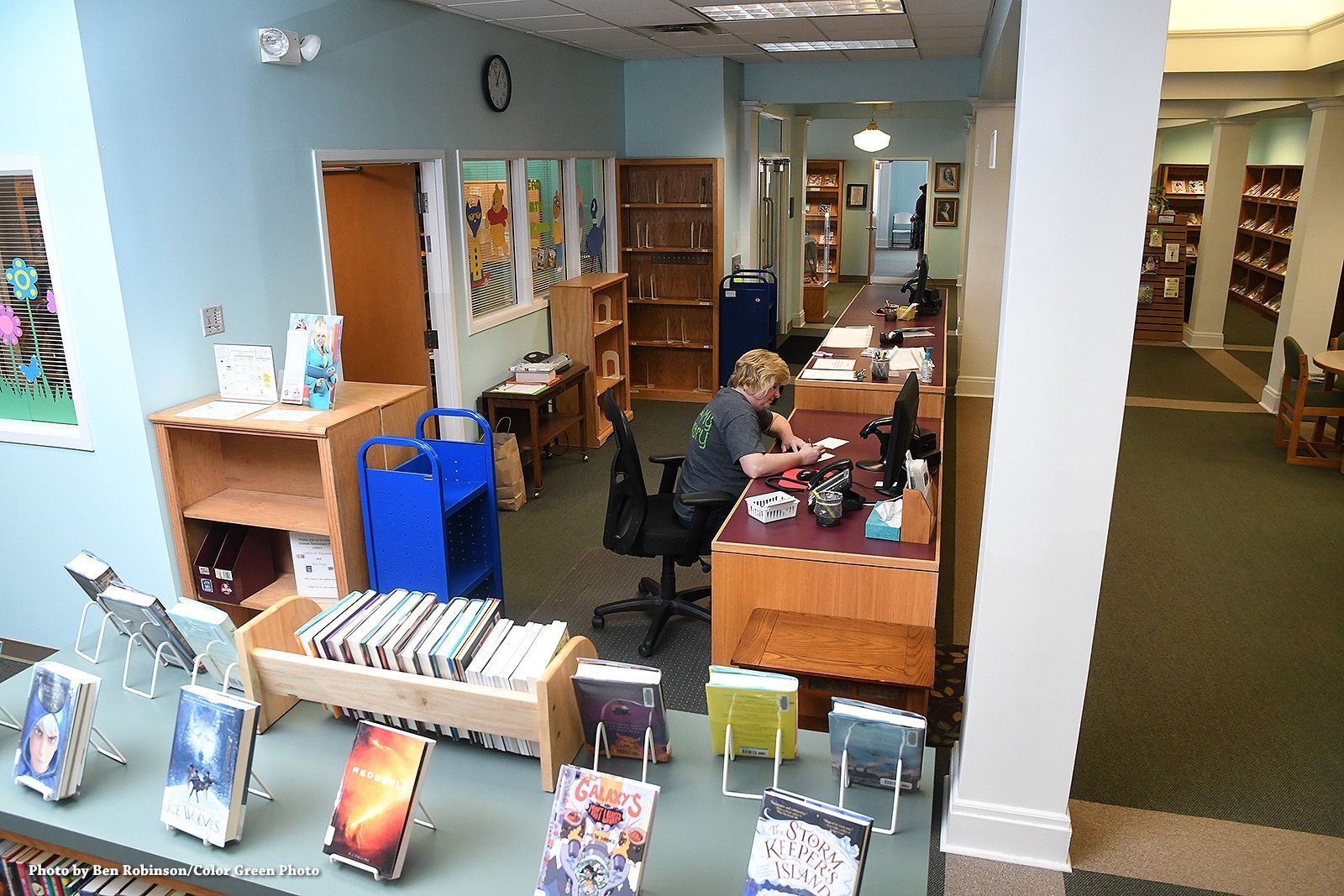 Library interior with book displays, check-out desk, and person working. Blue walls, green carpet.