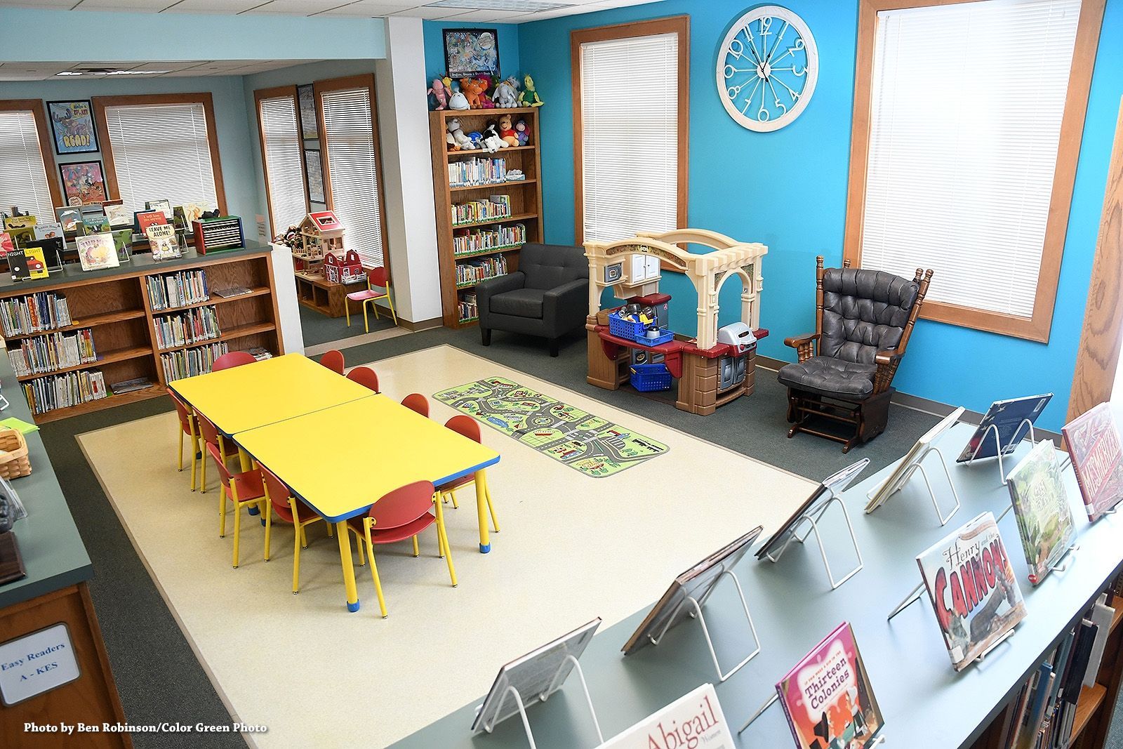 Children's library: bright blue walls, yellow table, chairs, bookshelves, playhouse, clock, and two chairs.