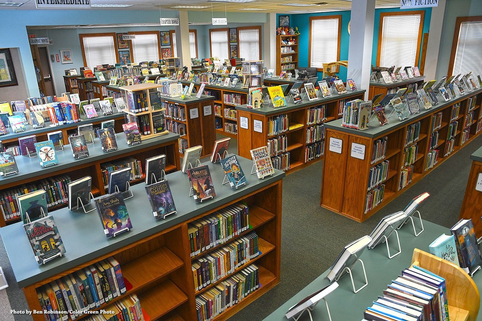 Library interior with bookshelves and tables displaying books. Sunlight streams in.