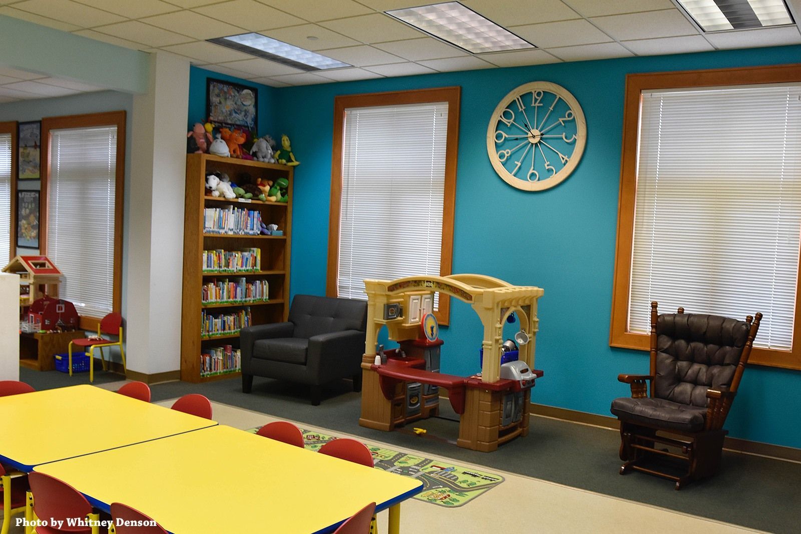 Children's library corner: turquoise wall, bookshelves, yellow table, playhouse, rocking chair, and wagon wheel decor.