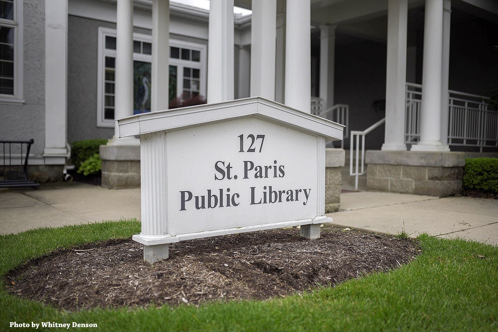 White sign in front of St. Paris Public Library, 127, on a green lawn.