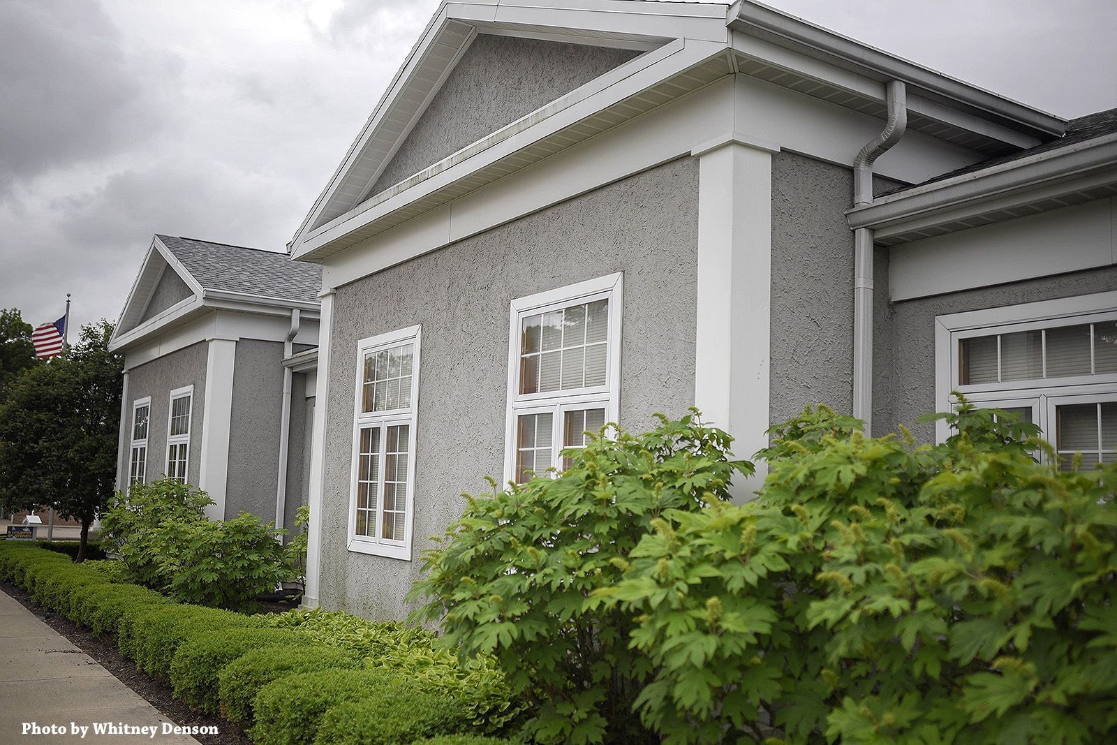 Gray stucco building with white trim, windows, and greenery in front.