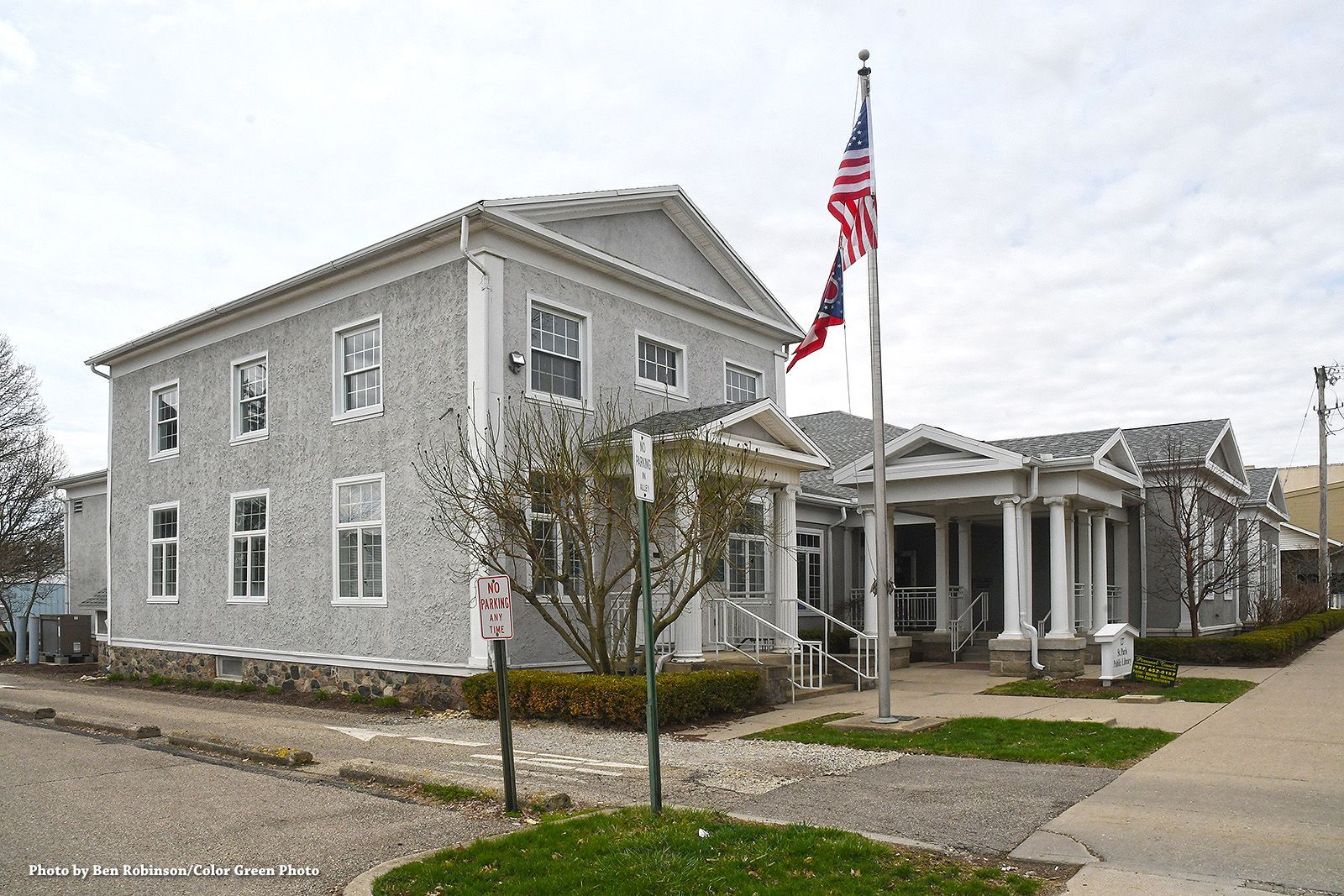 Two-story light gray building with white trim, flag pole, green lawn, sidewalk, and cloudy sky.