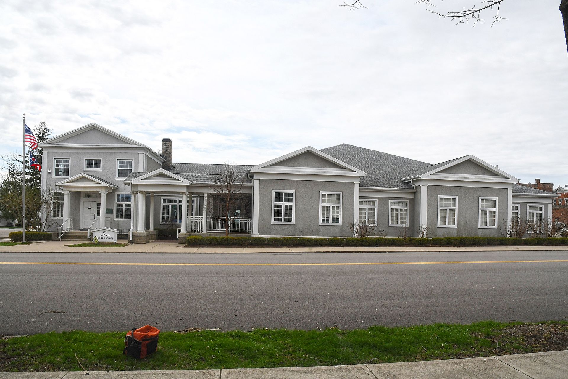 Gray building with several sections, pillars, and a flag. Sidewalk and street in front.