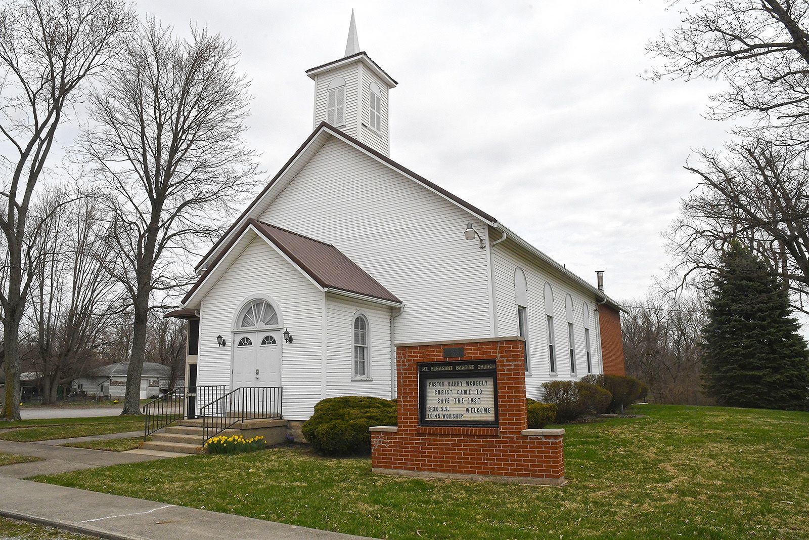 White church building with steeple, brown roof, and brick sign in front, cloudy day.