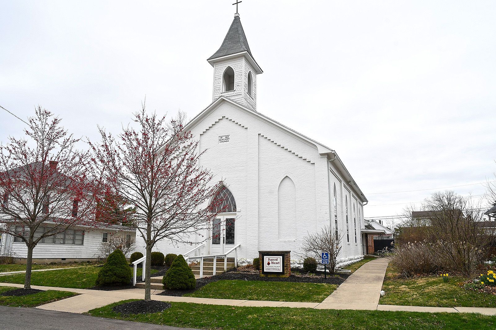 White church with steeple and cross on a cloudy day, framed by trees and a sidewalk.