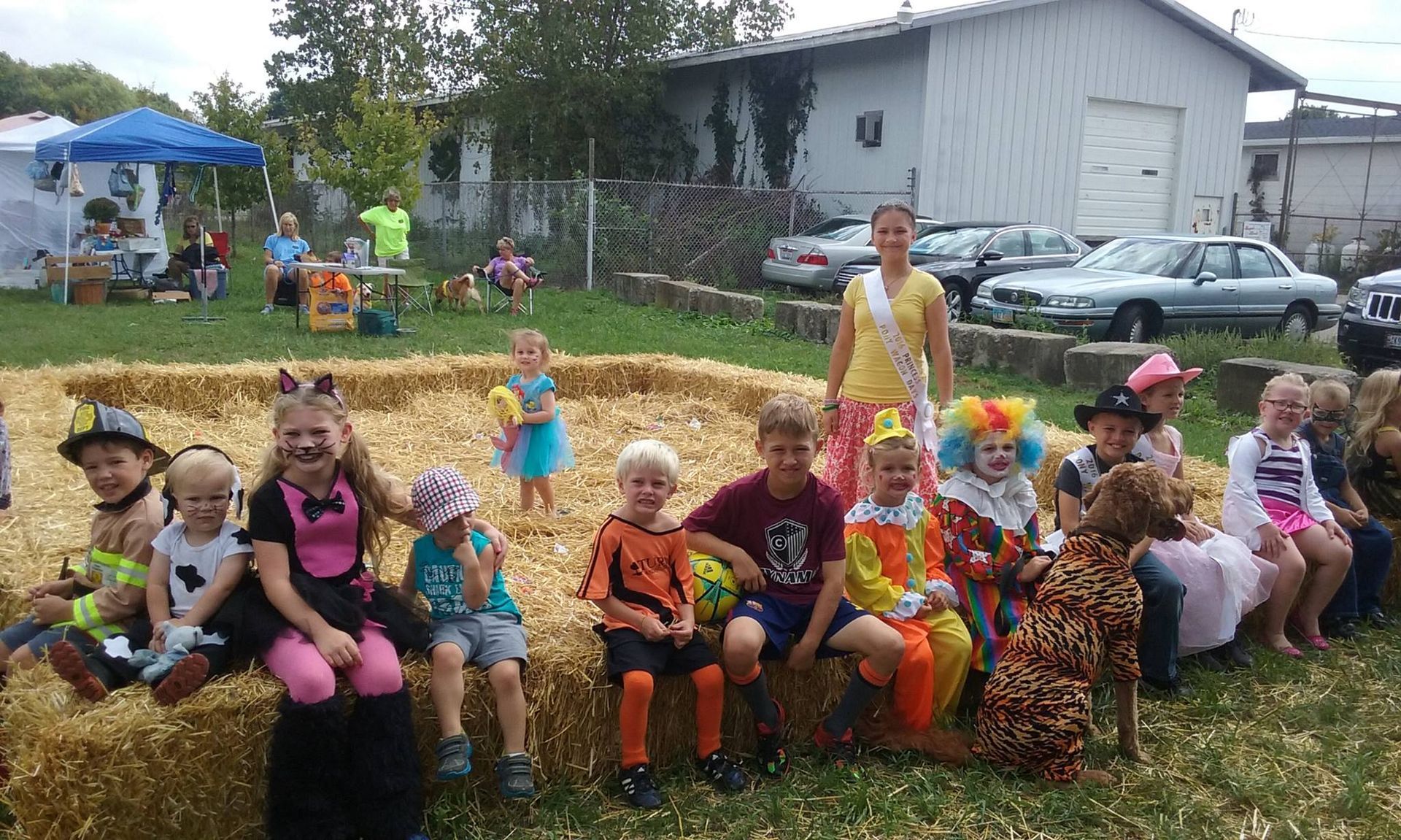 Children in costumes sitting on hay bales at an outdoor event. A woman with a sash stands nearby.