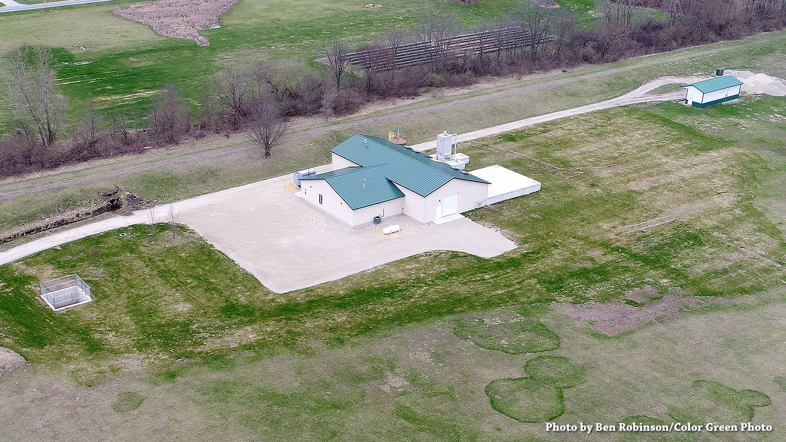 Aerial view of a light-colored building with a green roof next to a gravel area and a long pathway.