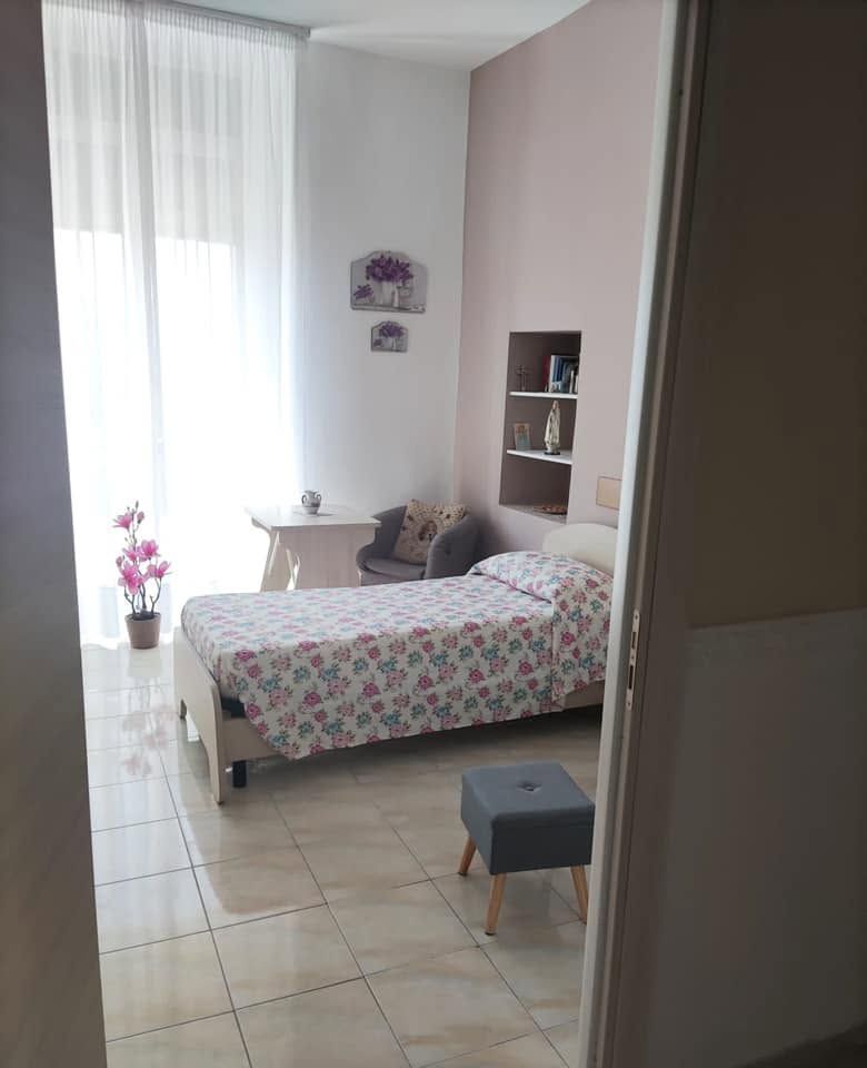 Camera da letto luminosa con copriletto floreale, pareti bianche, pavimento piastrellato e luce del sole che filtra attraverso tende trasparenti.