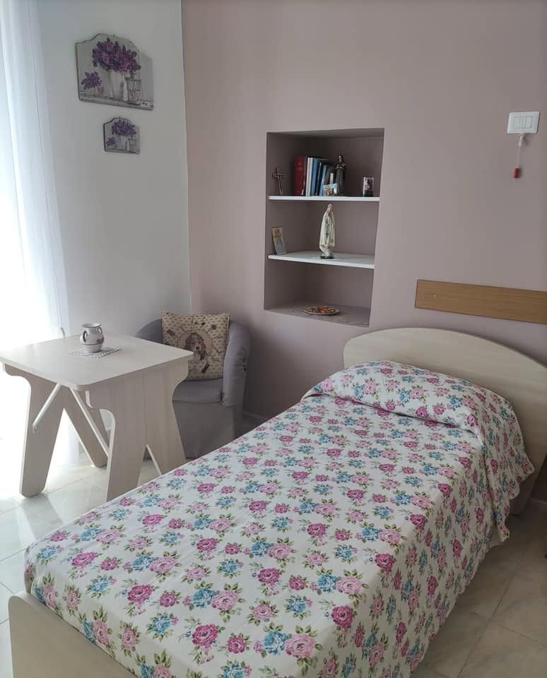 Piccola camera da letto con letto singolo, copriletto a fiori, mensola a muro, comodino e sedia vicino alla finestra.