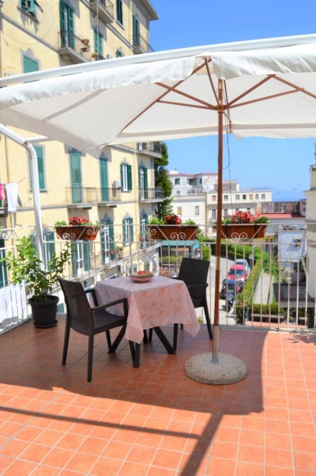 Terrazza panoramica soleggiata con tavolo, sedie e grande ombrellone bianco, con vista sugli edifici e sulla strada cittadina.