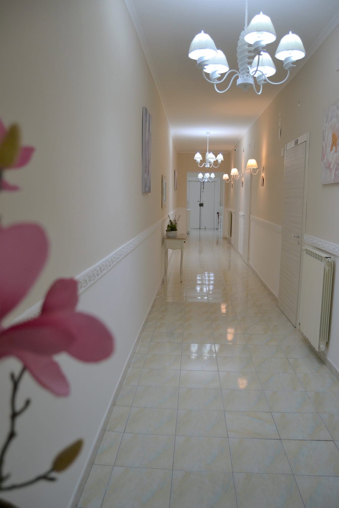 Elegante corridoio color crema con pavimento in marmo lucido, lampadari bianchi e fiori rosa in primo piano.